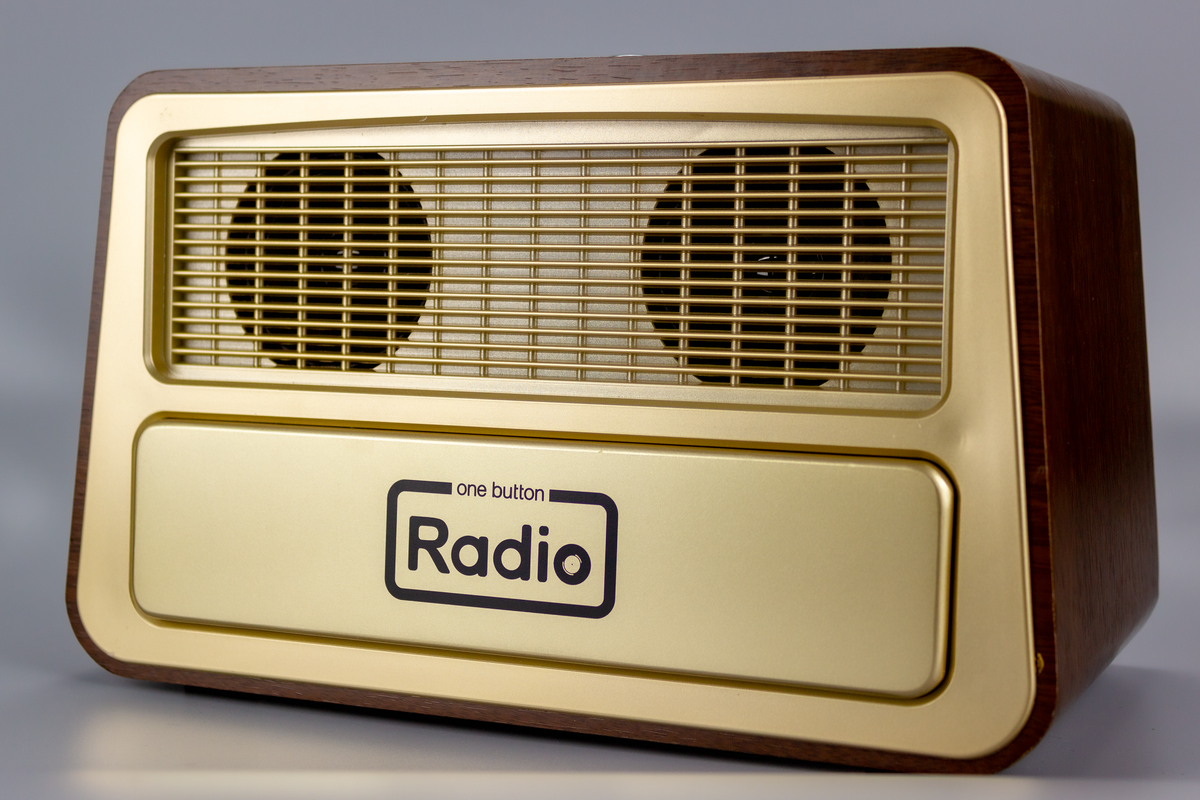 The radio rectangular in shape and gold-coloured with dark brown sides and front border.  The front of the radio is divided equallly into two horizontal rectangular halves.  The top half is shallowly indented and within this sit two equidistant round flat speakers. They are overlaid with a gold-coloured plastic rectangular grid. The bottom half has a l