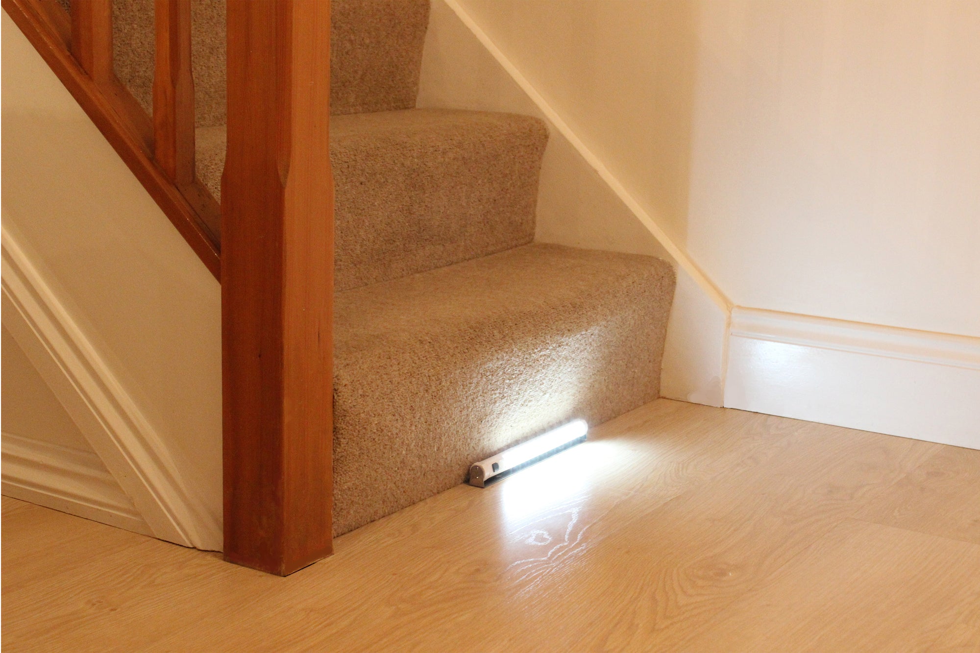 In the middle of the picture, the bottom of a flight of beige-carpeted stairs with wooden bannisters is visible. The setting is a domestic interior with white wall and laminate flooring. Only the first three stairs are shown and they are seen from a side/front angle.  A tubular illuminated light is positioned on the laminate flooring so that it sits firmly against the middle of the bottom stair.