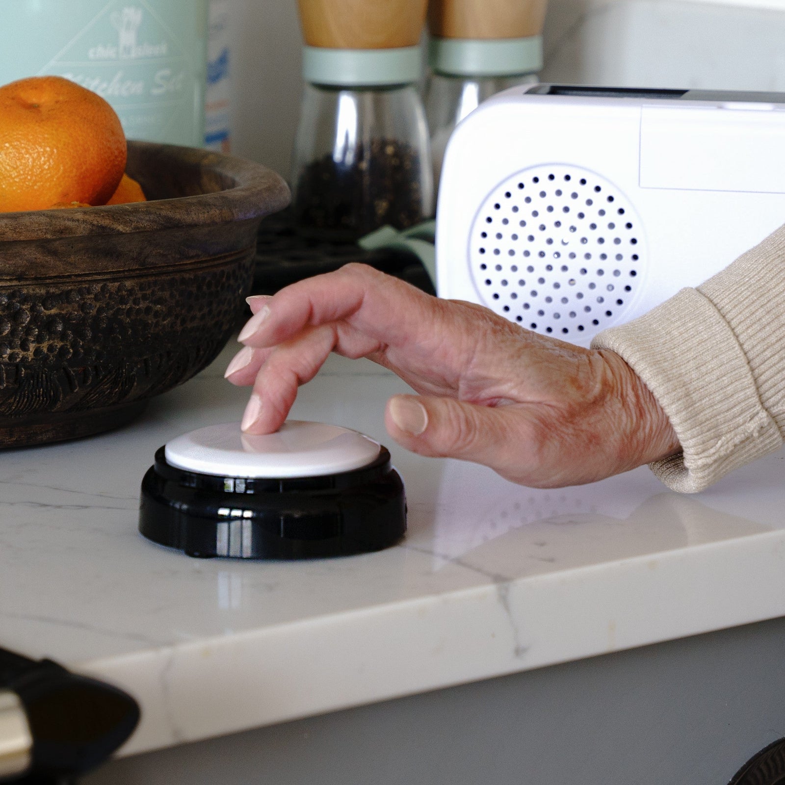 Hand pressing a black and white button on a kitchen counter with a white radio and bowl of oranges in the background.