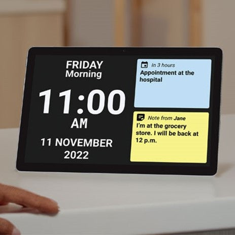 A black digital tablet displaying the time, date and sticky note messages, sat on a white kitchen top with someone's hand resting nearby.