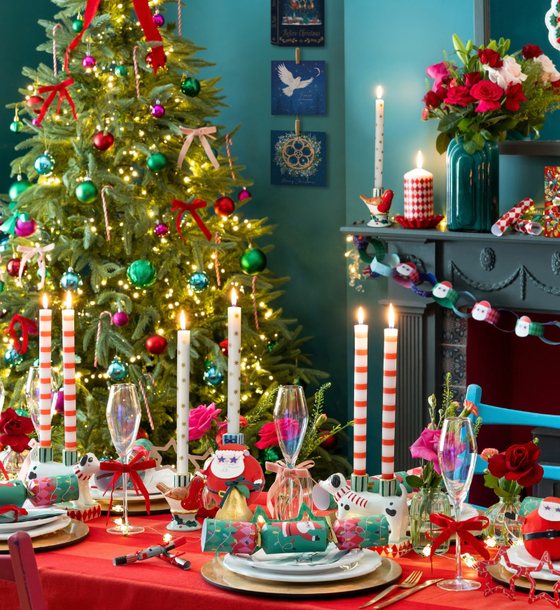 Festive Christmas scene with a lit up Christmas tree with bright coloured baubles & bows. Fireplace in the background with a santa paper chain and candles on top. Bright coloured table setting with santa crackers and tall patterned dinner candles.