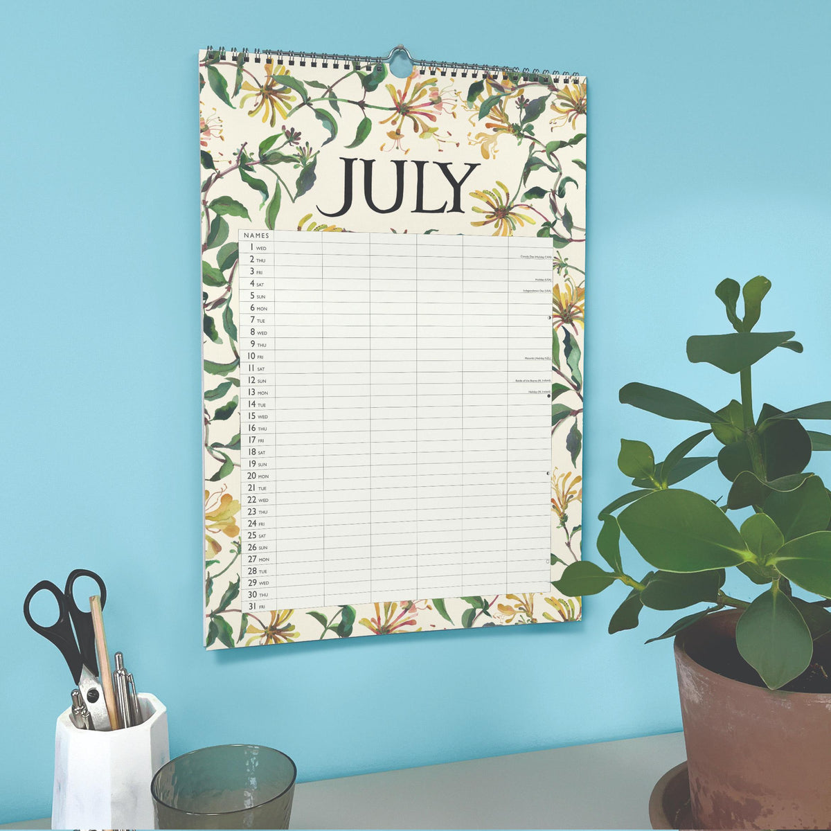 Floral-patterned calendar for July on a blue wall with stationery and a plant.