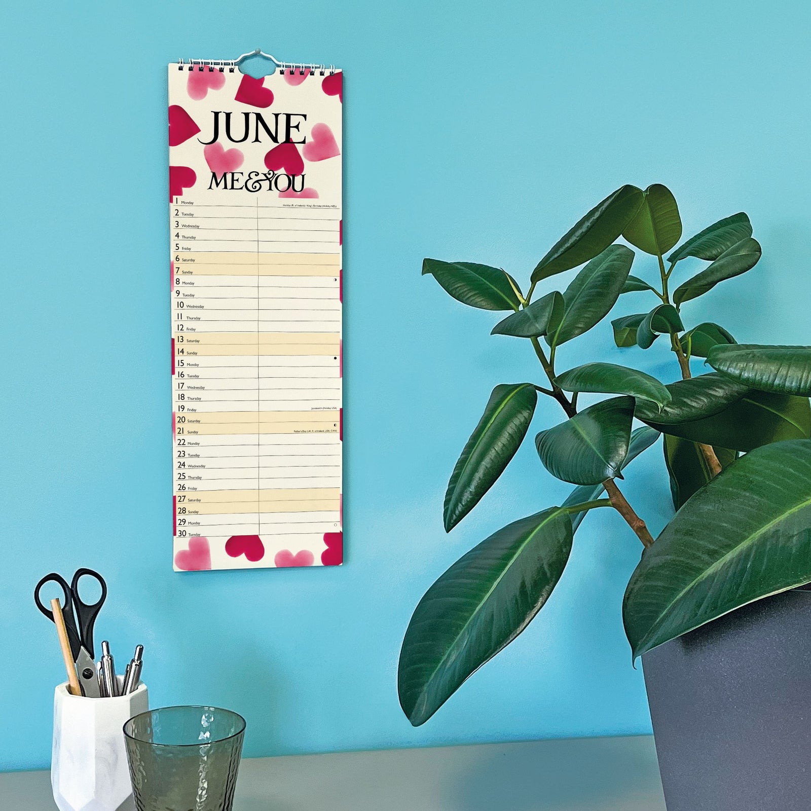 Vertical calendar for June with floral design, placed on a blue wall next to a potted plant.