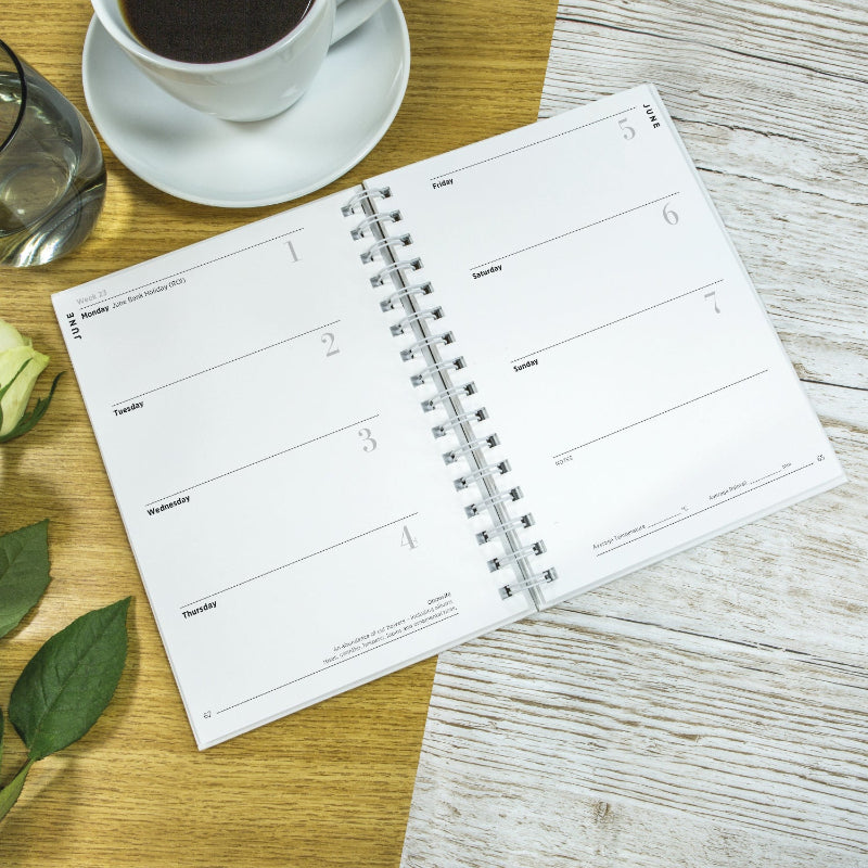 Open planner on a wooden table with a cup of coffee and green leaves.