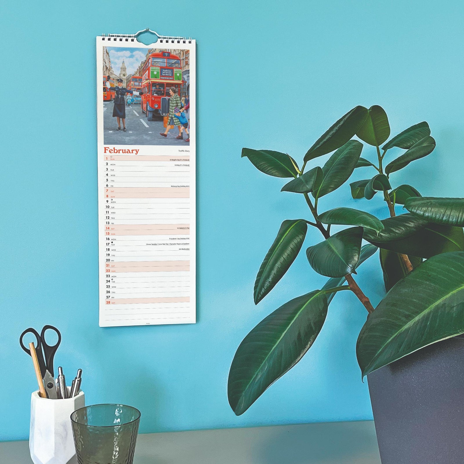 Wall calendar on a blue wall with a plant and stationery items below.