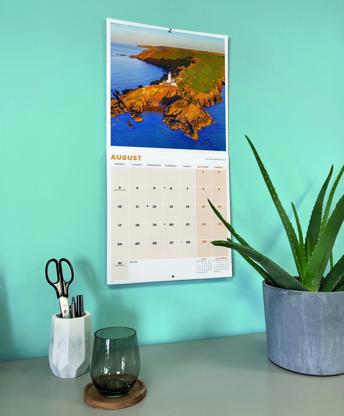Calendar with a scenic design on a teal wall, next to a plant and desk items.