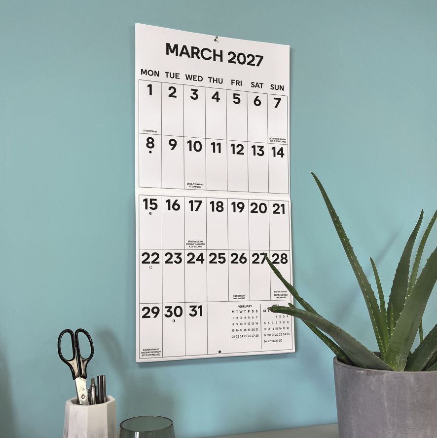 A white March 2027 wall calendar hanging on a light blue wall above a desk with a potted aloe plant, a glass, and a pencil holder.