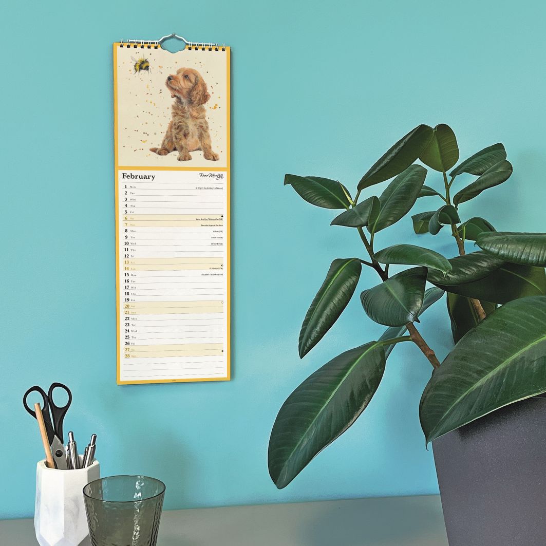 A vertical wall calendar page featuring a puppy illustration and a monthly planner layout, hanging on a turquoise wall in a styled room scene with a potted plant and desk accessories.
