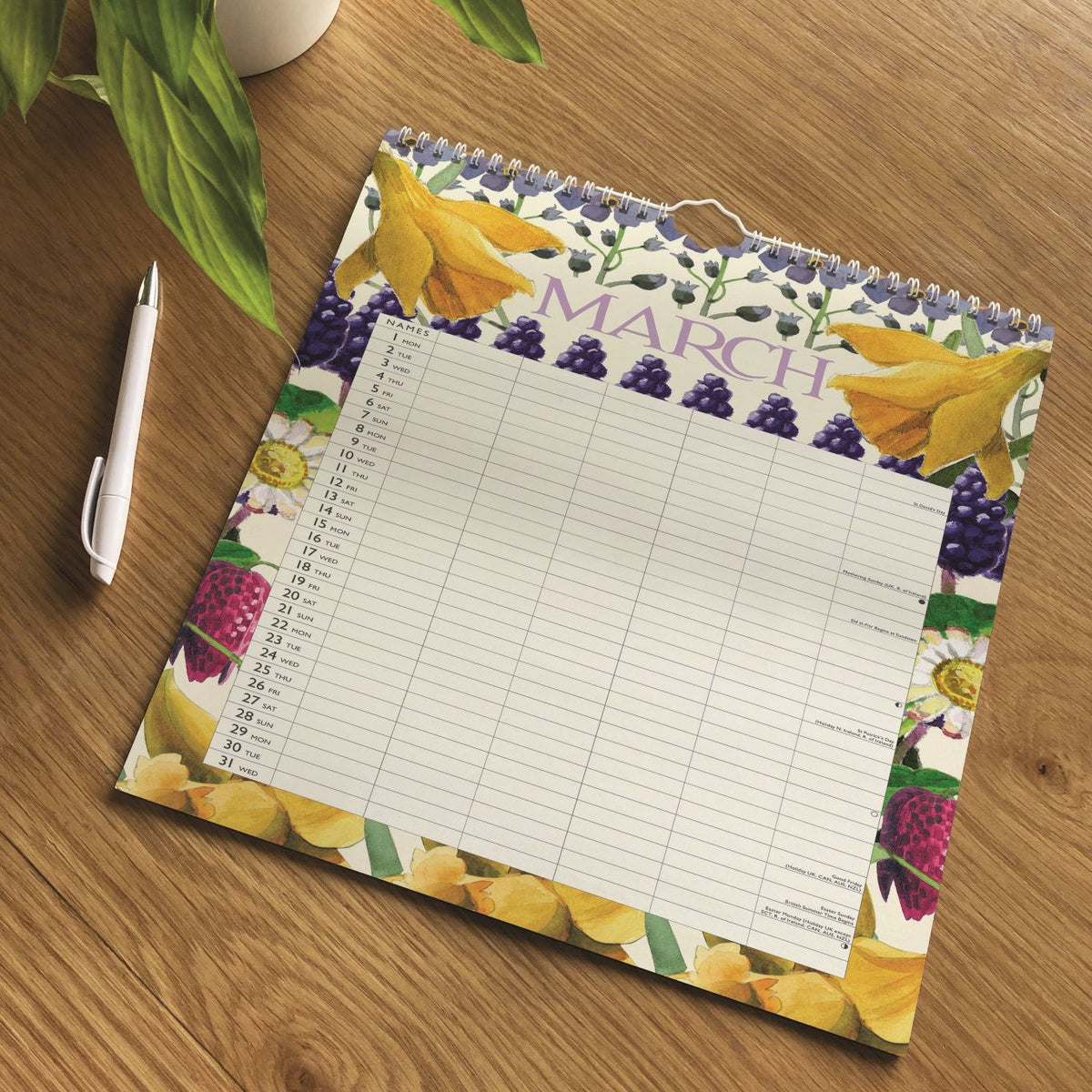 A spiral-bound March calendar page with a fruit and flower border design, placed on a wooden tabletop next to a white pen. The calendar has a lined writing area in the center and the word MARCH printed across the top.