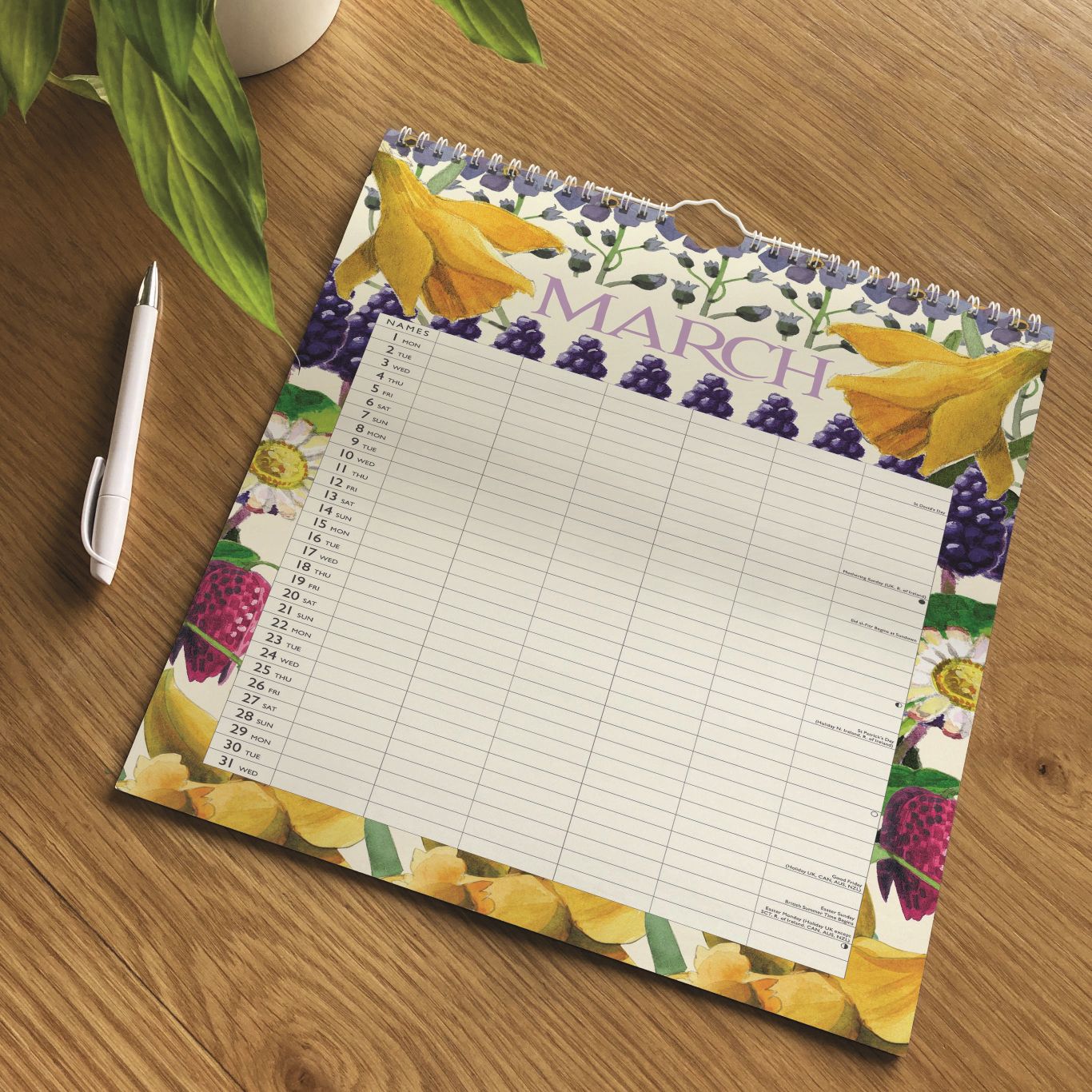 A spiral-bound March calendar page with a fruit and flower border design, placed on a wooden tabletop next to a white pen. The calendar has a lined writing area in the center and the word MARCH printed across the top.