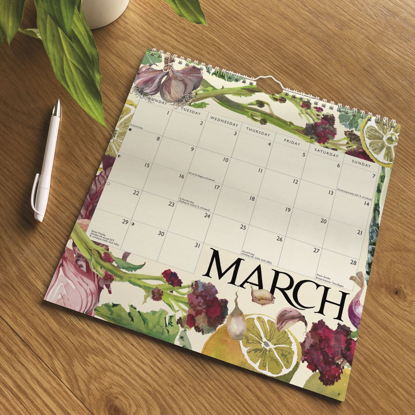 A March desk calendar with a botanical and food-themed border featuring garlic, onions, lemons, and grapes, displayed on a wooden tabletop next to a white pen and a plant.