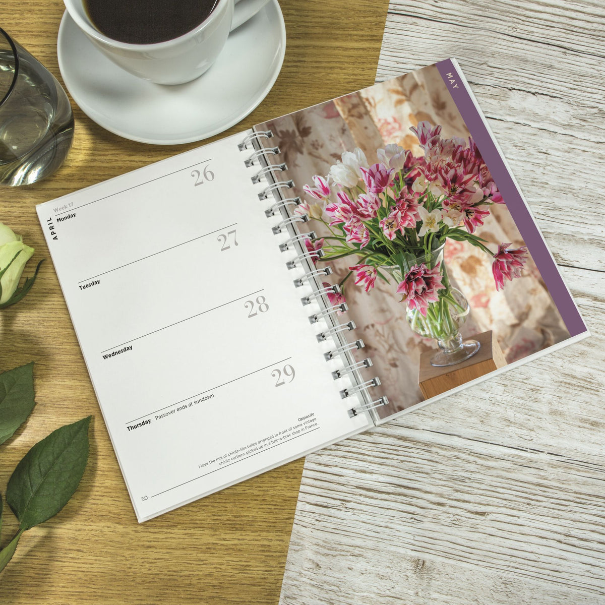 An open spiral-bound weekly planner or calendar lies on a wooden surface, showing printed weekday layouts and a floral photo on the right page. A white coffee cup and saucer, a glass, and green leaves are partially visible around it.