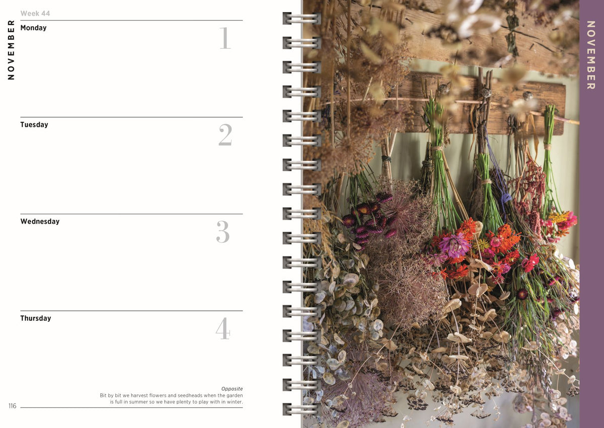 A spiral-bound planner or notebook shown open to a weekly layout page on the left and a floral cover or interior spread on the right, featuring dried flowers and a vertical November label.