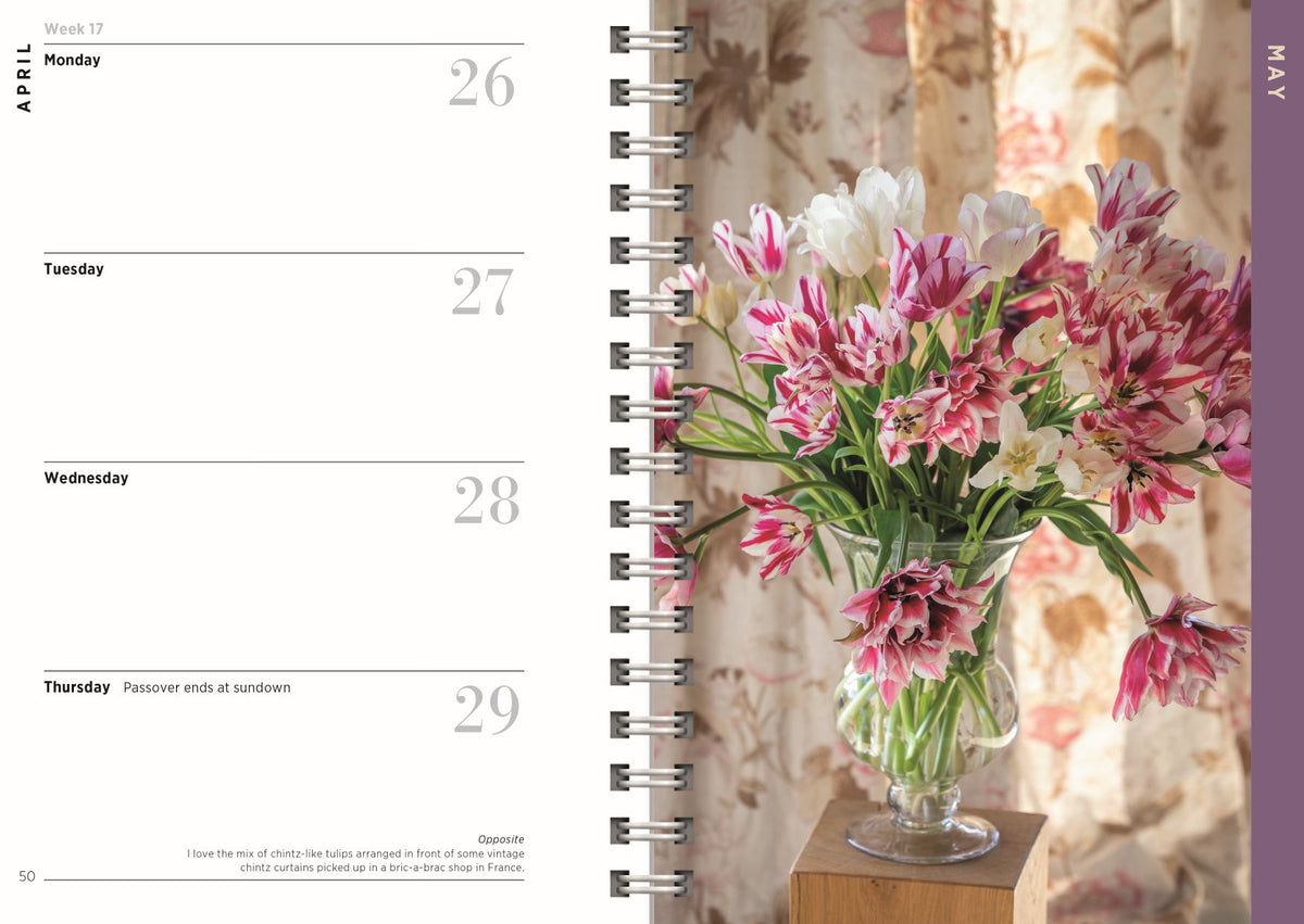 A spiral-bound planner or calendar spread showing April weekdays on the left and a floral scene on the right. The right page features pink and white flowers in a clear glass vase on a wooden surface against a patterned curtain background.