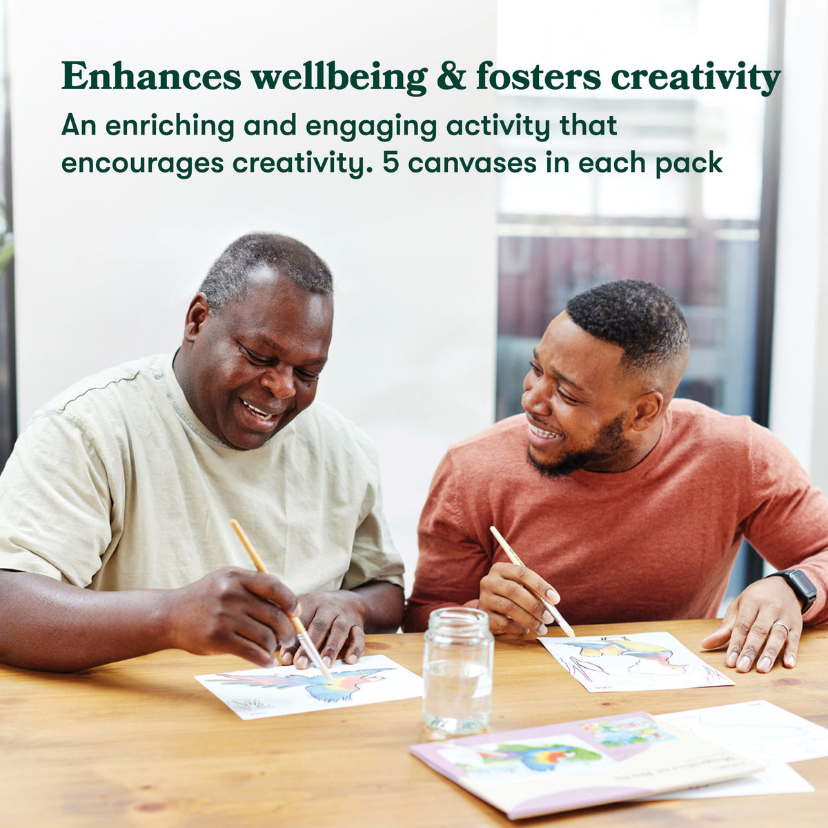 Two men sitting at a table with art supplies, smiling and engaged in an activity.