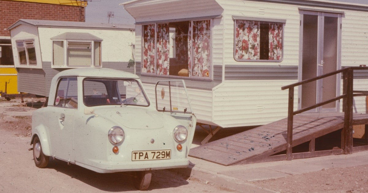 Small vintage car parked in front of a trailer home with floral curtains.