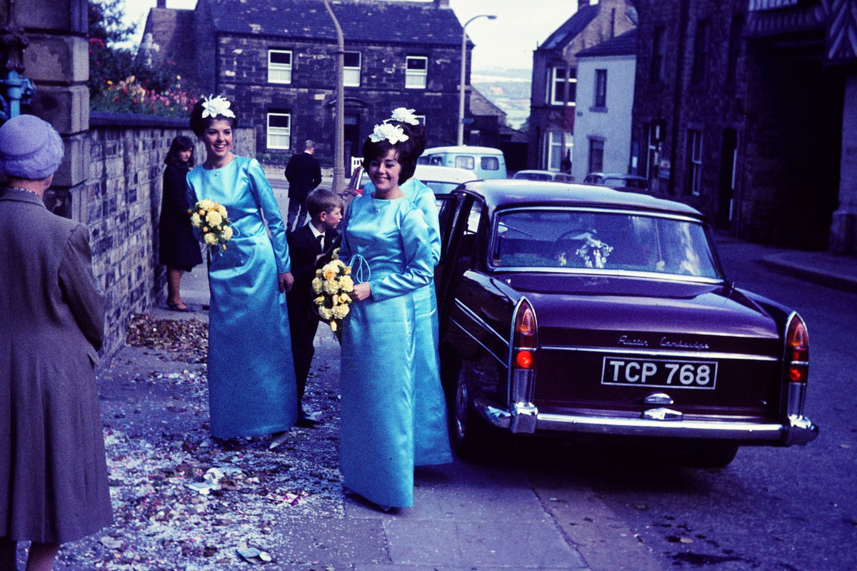 Three women in blue bridesmaid dresses with floral bouquets standing next to a burgundy car on a street.