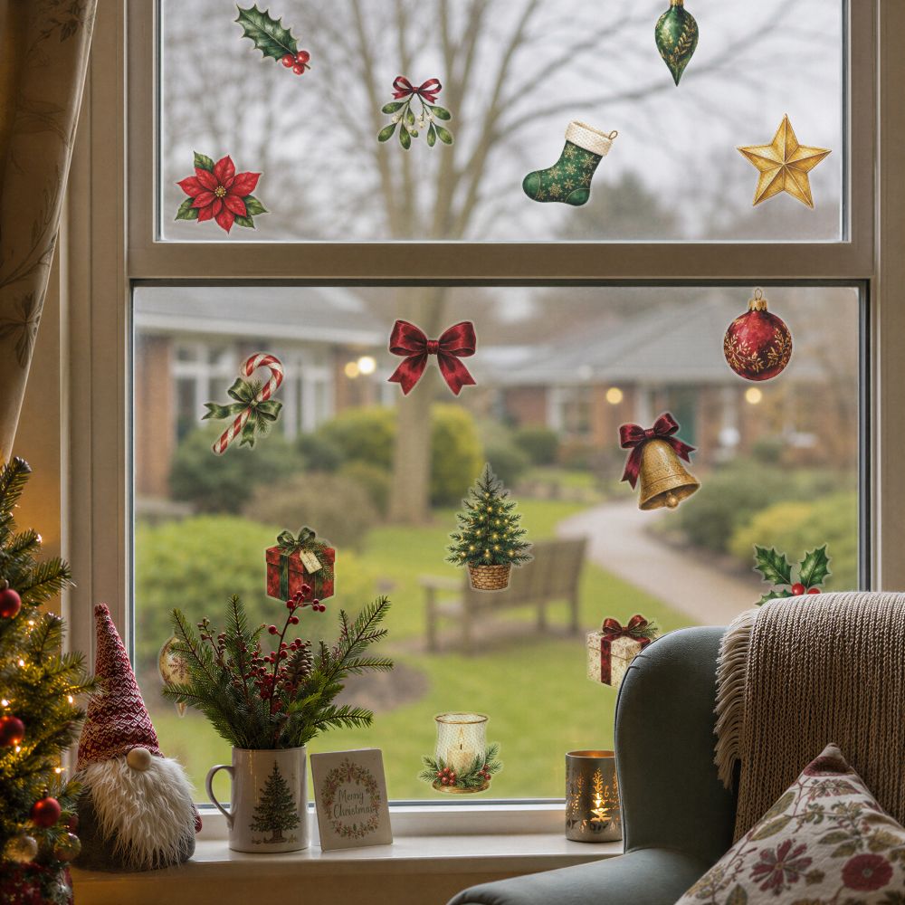 A set of Christmas window decals displayed on a glass windowpane, showing a cozy holiday scene with a window seat, a green armchair, a decorative gnome, and a garden view outside.