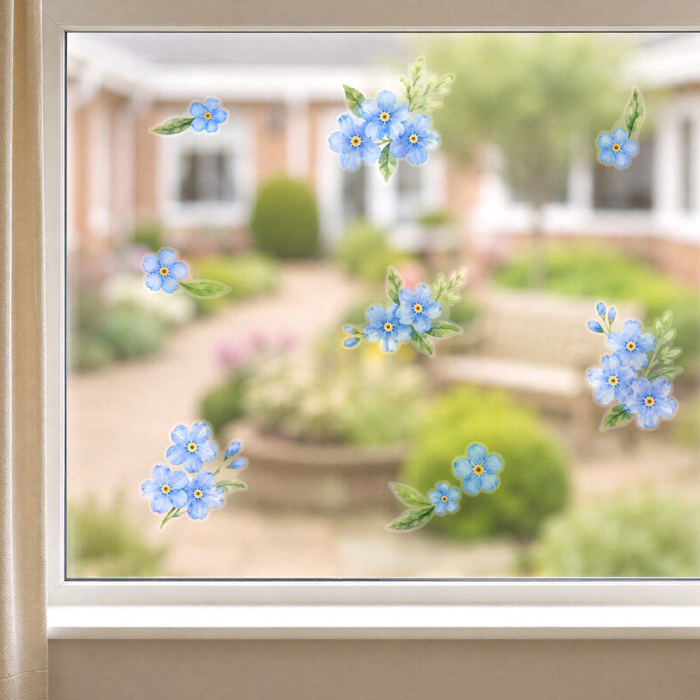 Blue forget-me-not flower window decals with green leaves and yellow centers applied to a glass window, with a blurred garden background visible behind them.