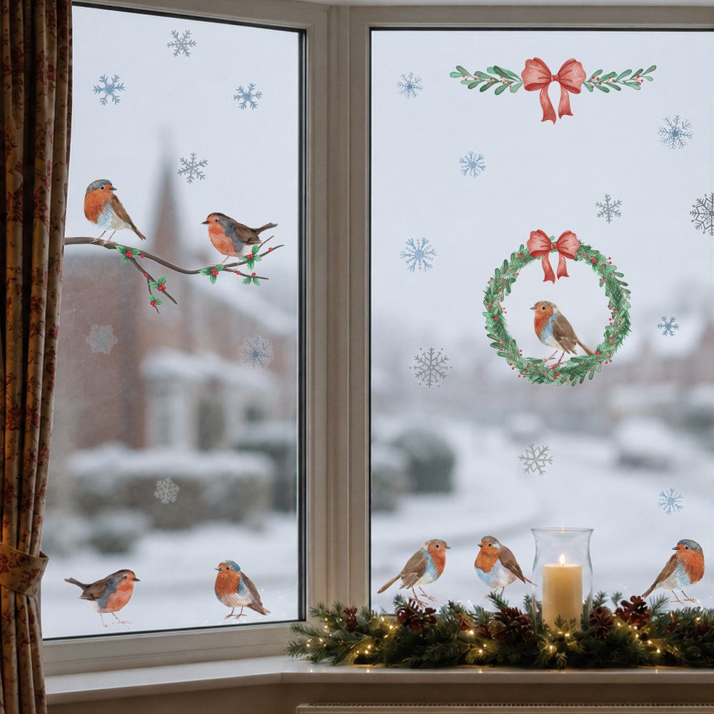 Christmas-themed window decals featuring robin birds, a wreath, snowflakes, and a red bow, shown applied to a snowy window with a decorated window sill below.