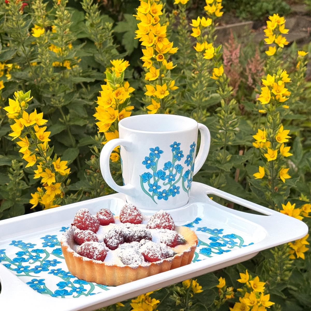 White mug with floral design and a tart on a tray against a natural background with yellow flowers.