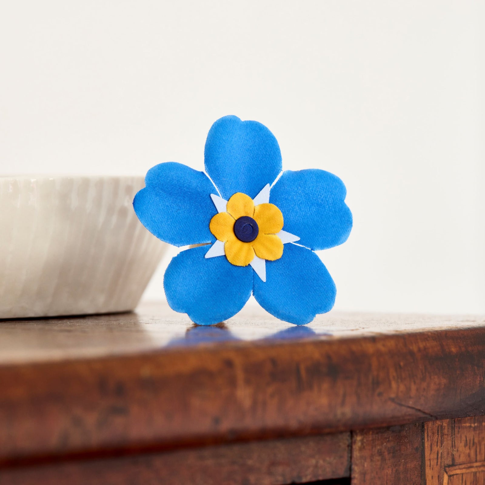 Blue and yellow forget me not appeal fabric flower on a wooden surface with a white background