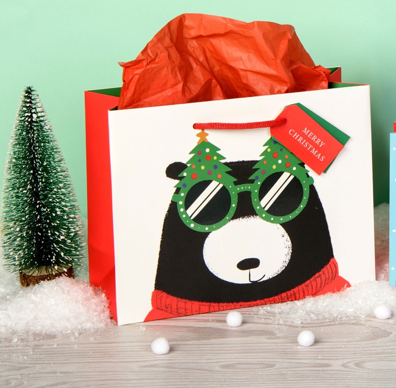 A white Christmas-themed gift bag with a cartoon bear wearing green party glasses and a red sweater, filled with bright orange tissue paper. The bag has a red handle and a red 'Merry Christmas' tag.