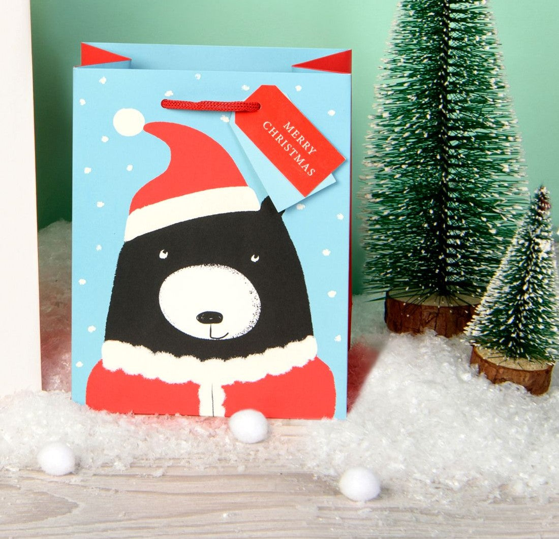 A light blue Christmas gift bag featuring a cartoon black bear wearing a red Santa hat and scarf, styled with artificial snow, small green bottle tree decorations, and a red 'Merry Christmas' tag.