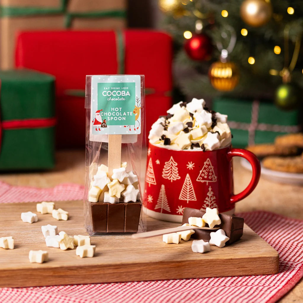 Hot chocolate spoon with star marshmallows in a packet, next to a festive mug with whipped cream and marshmallows on top.