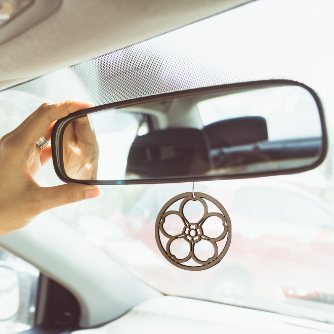 A wooden laser-cut forget-me-not decoration is hanging by a white string from a car's rear view mirror. The mirror is being adjusted by a person's hand and the rear of the car interior is visible in the mirror.