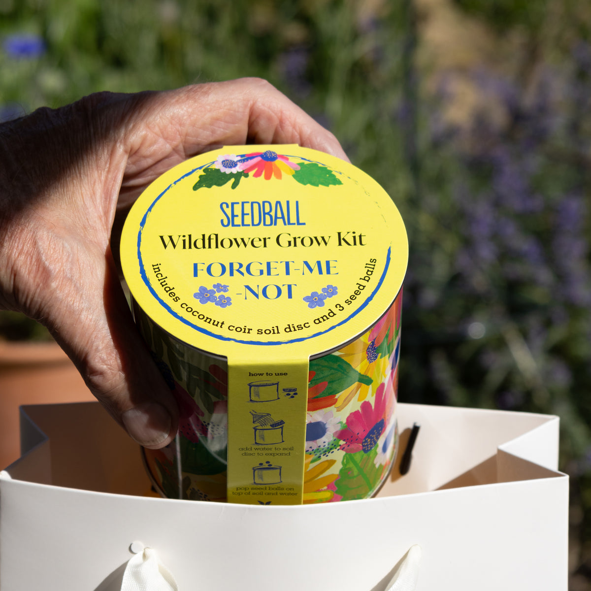 Someone's hand holding a floral decorated tin with the wording Seedball Wildflower Grow Kit Forget-me-not on the label telling you that the tin includes coconut coir soil disc and 3 seed balls.