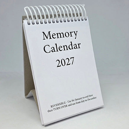 A spiral-bound 2027 desk calendar standing upright using the cardboard base as a small display stand, with a white cover and black text reading 'Memory Calendar 2027' on a light gray background.