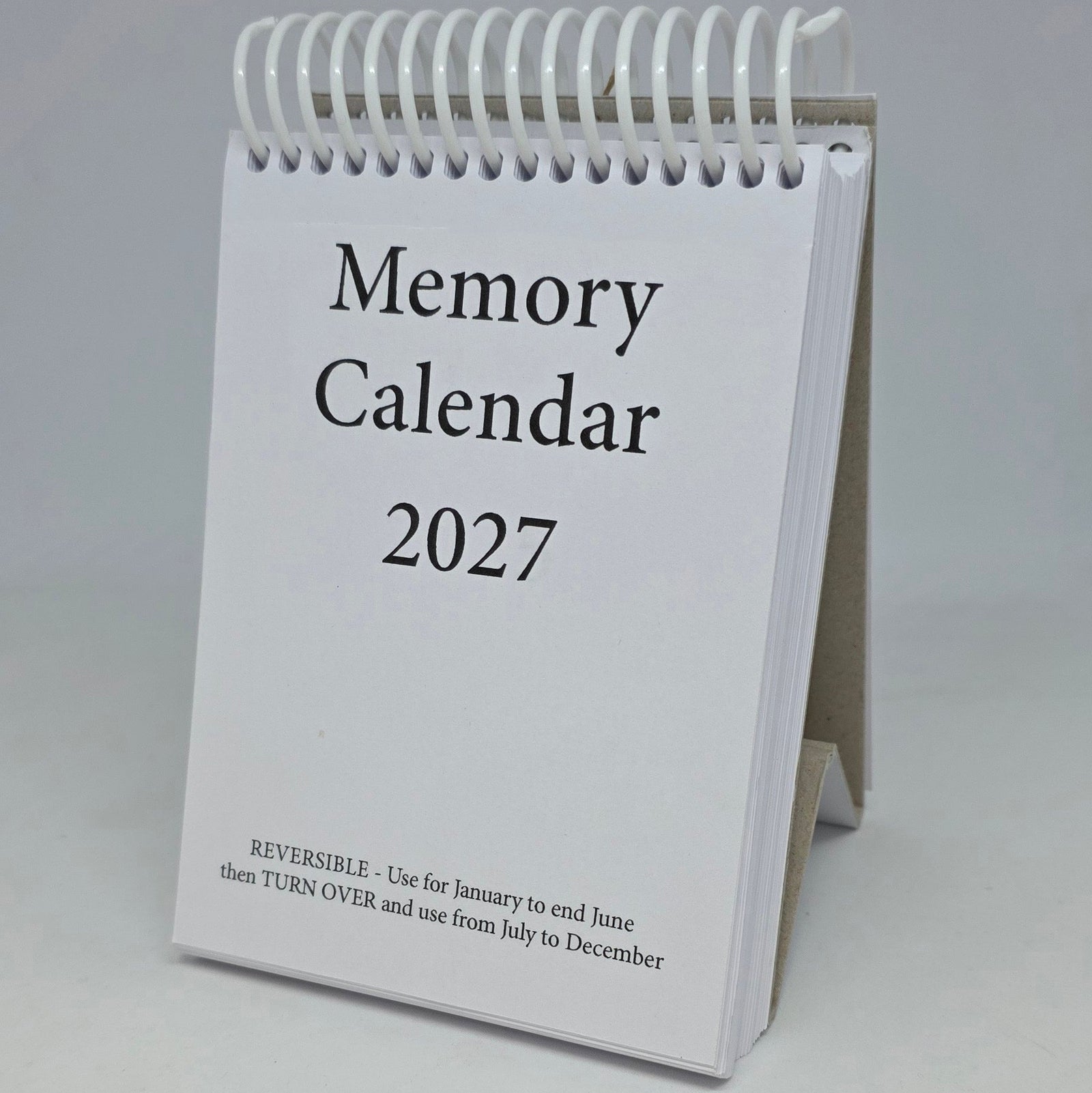 A spiral-bound 2027 desk calendar standing upright using the cardboard base as a small display stand, with a white cover and black text reading 'Memory Calendar 2027' on a light gray background.