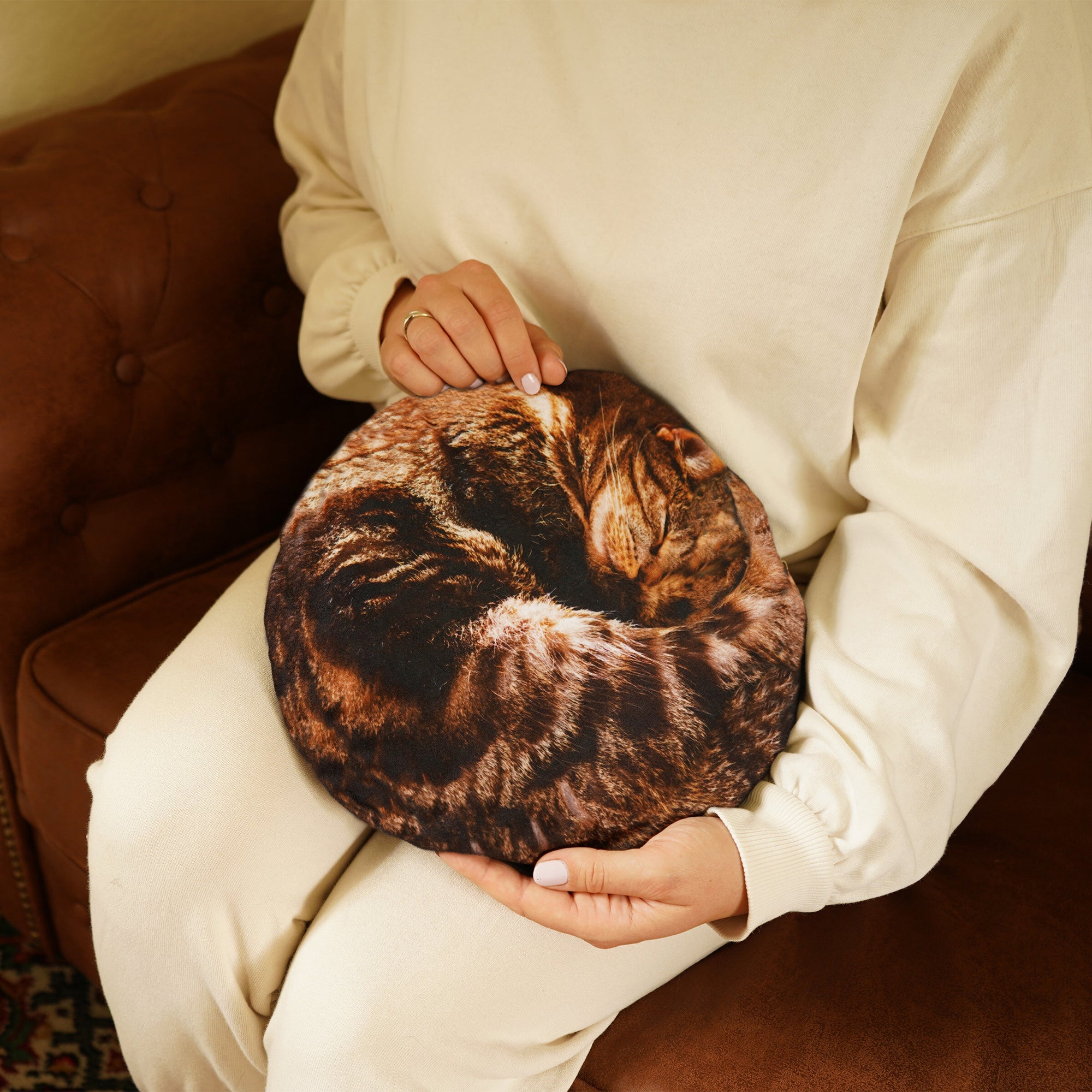 A lady sitting down with a hot water on her lap. The hot water bottle is round and has a sleeping cat design on it.