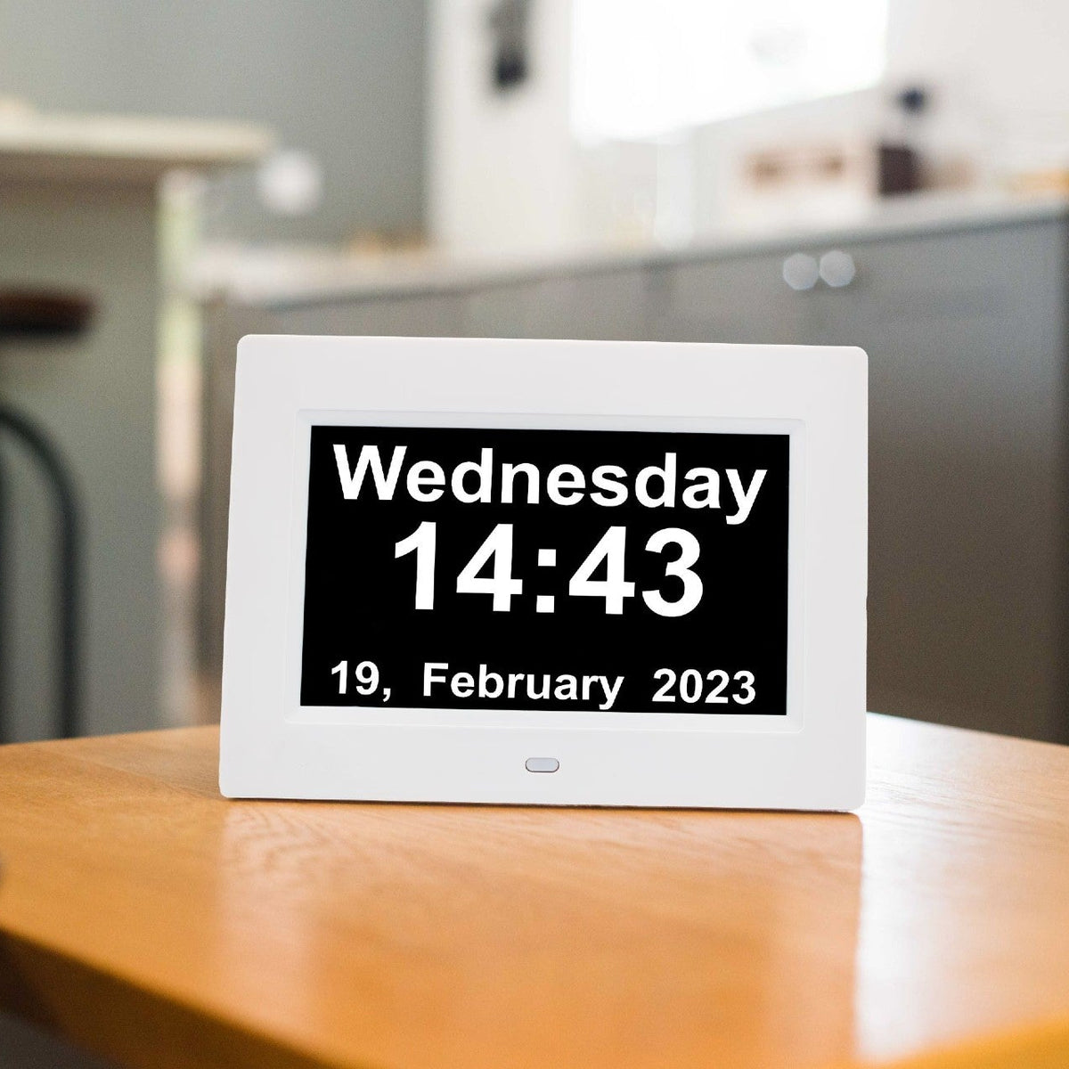 A white digital clock displaying 'Wednesday 14:43 19, February 2023' on a wooden table in a blurred kitchen setting.