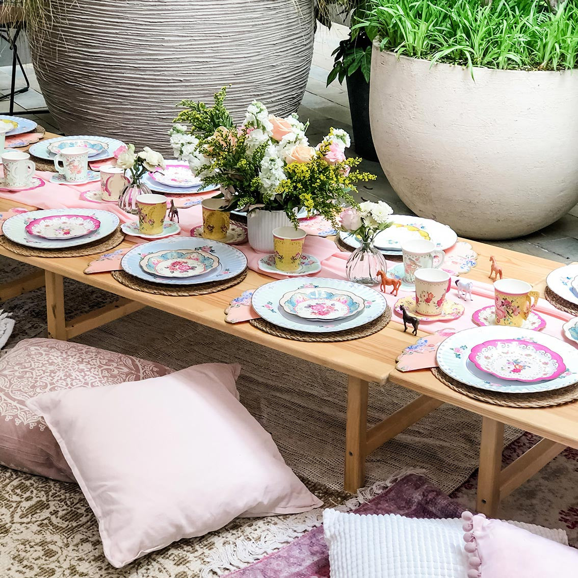 Garden table set up with cups, plates and flowers and cushions on the floor