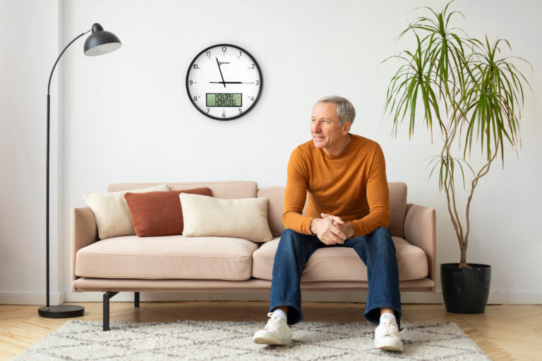 Large white analogue clock with digital display on a wall with a man sat on a sofa infront of it.