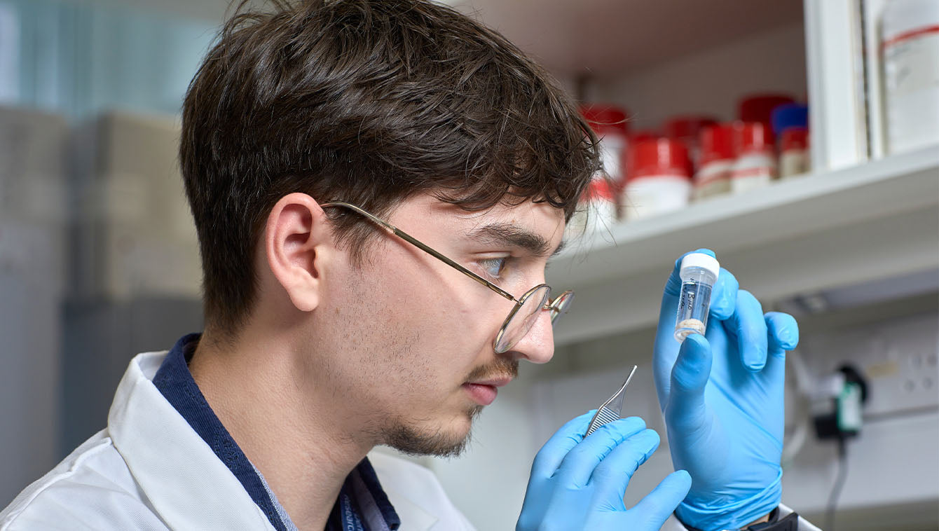 Person in a lab coat holding a small container and pipette in a laboratory setting