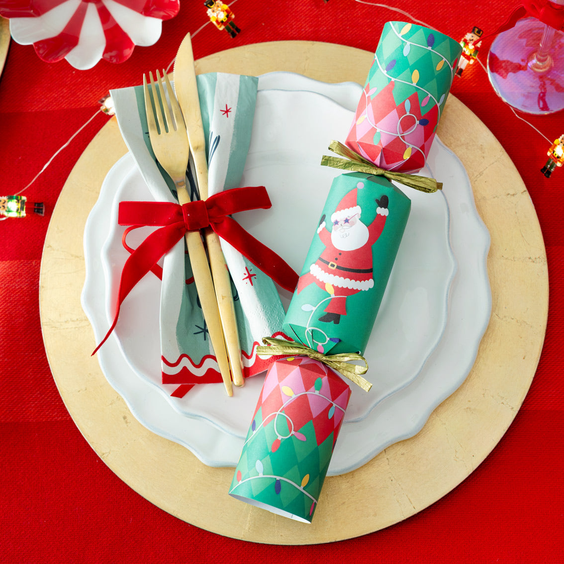 Christmas-themed crackers on a plate with cutlery and napkins on a red tablecloth.