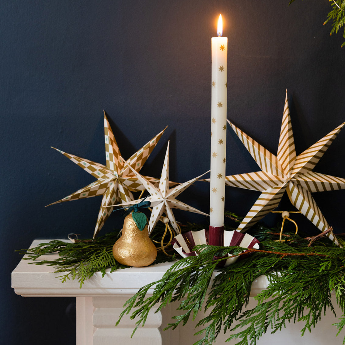 Decorative candle with gold stars on a white base, surrounded by star-shaped ornaments and greenery against a dark background.