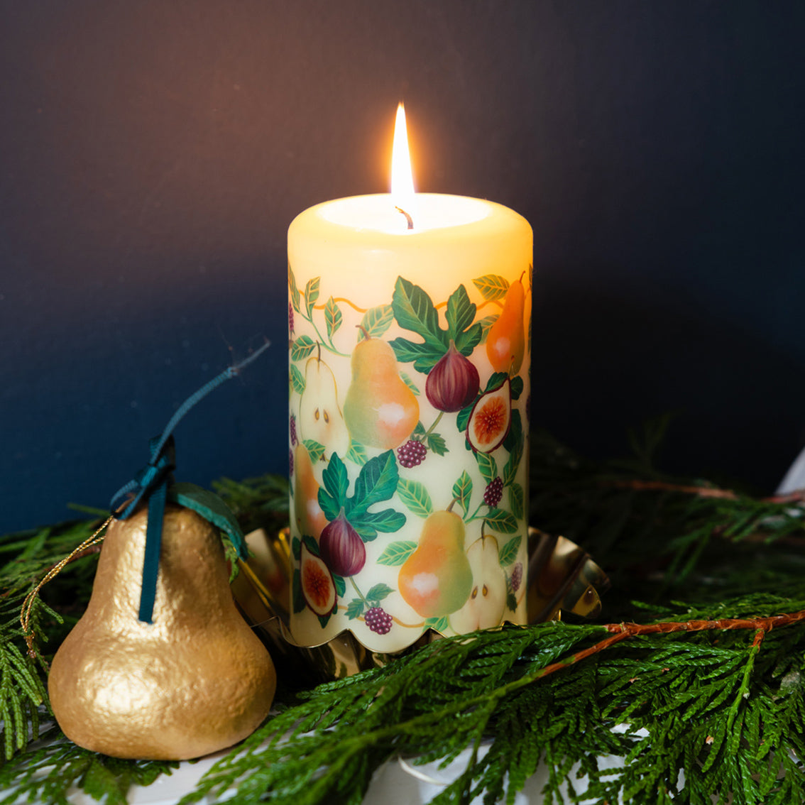 Decorative gold pear next to a candle with fruit design on a bed of greenery against a dark background