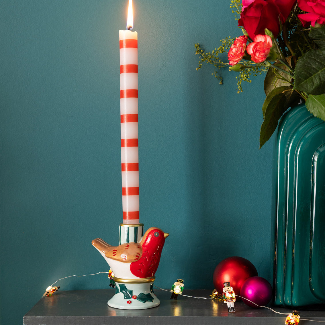 Decorative candle holder with a striped candle, bird design, and Christmas ornaments on a teal background.