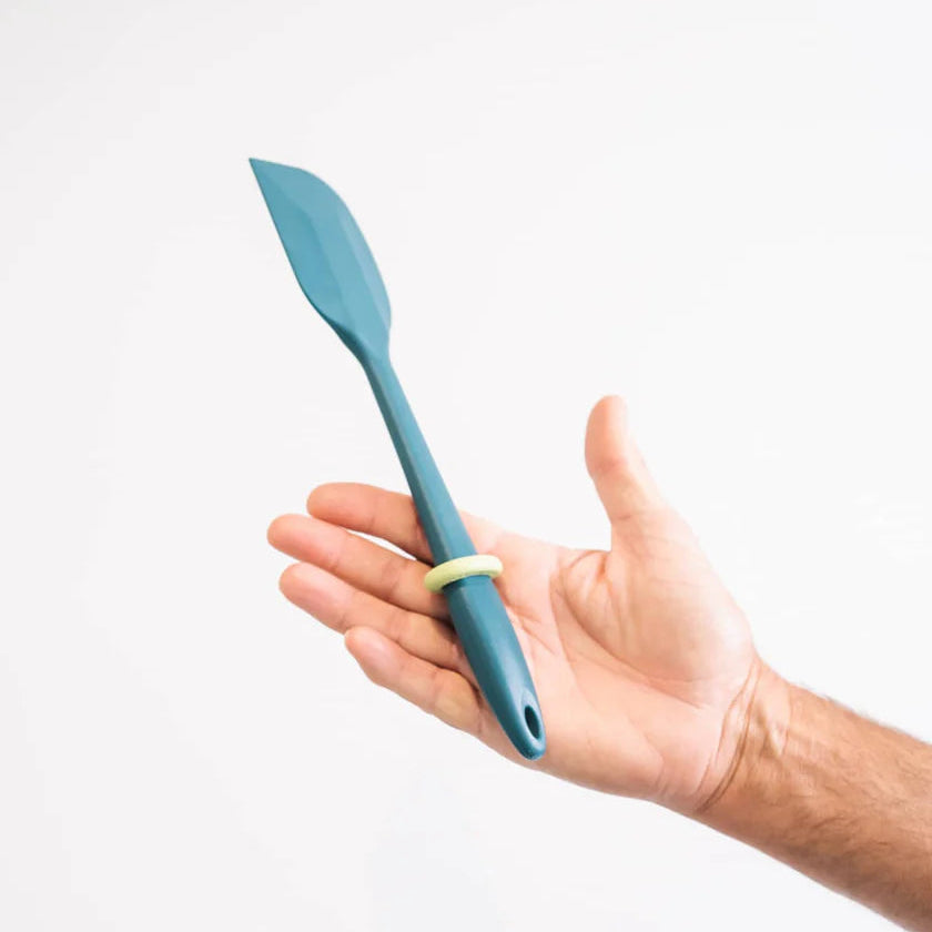 Hand holding a blue spatula with a green grip toggle against a white background