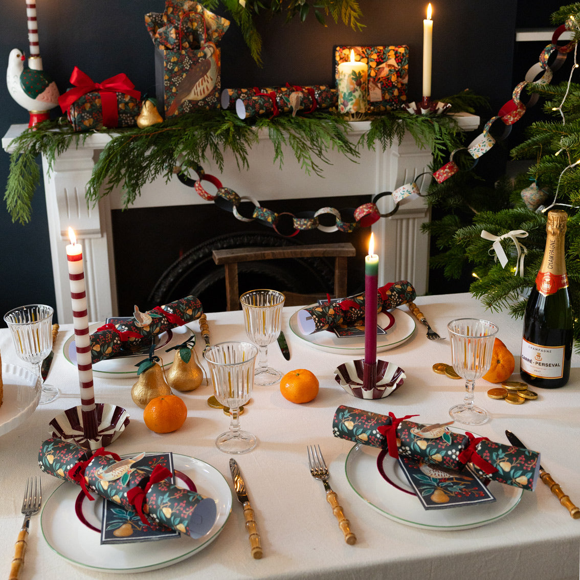 Decorated Christmas table with crackers, napkins, candles and a bottle of champagne in front of a fireplace.