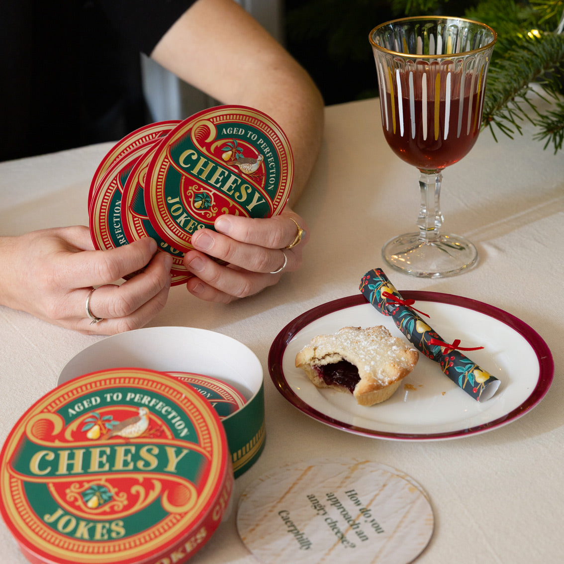 Person holding 'Cheesy Jokes' coasters with a festive setting including a glass of red drink and a mince pie.