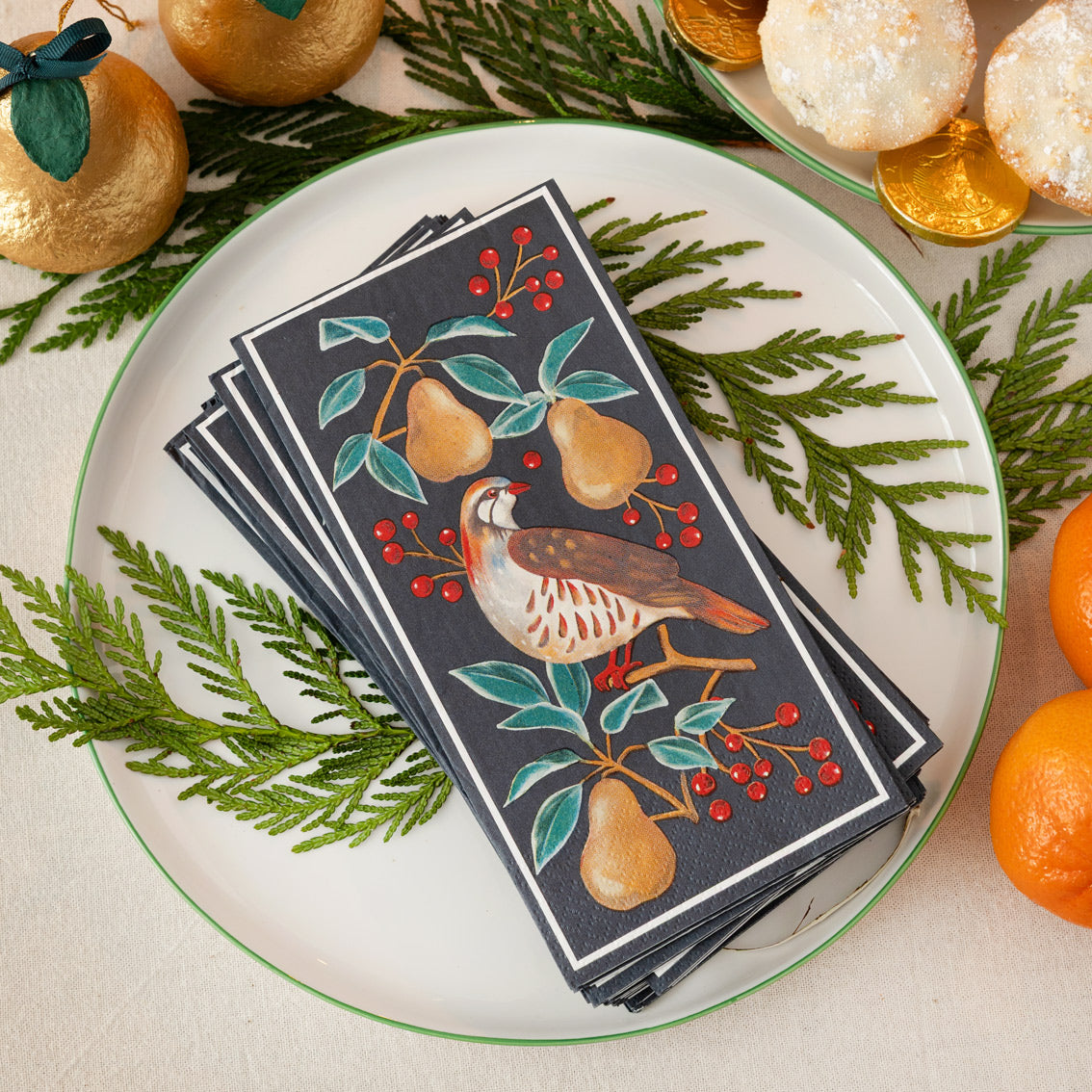Stack of decorative napkins with bird and pear design on a plate with greenery and oranges.