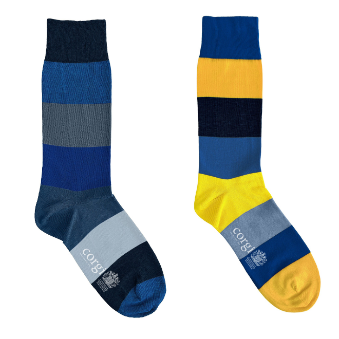 Two crew socks shown side by side on a plain white background. One sock has horizontal stripes in navy, blue, gray, and light blue with a navy toe and heel. Brand name Corgi and the King's royal warrant are on the sock sole.