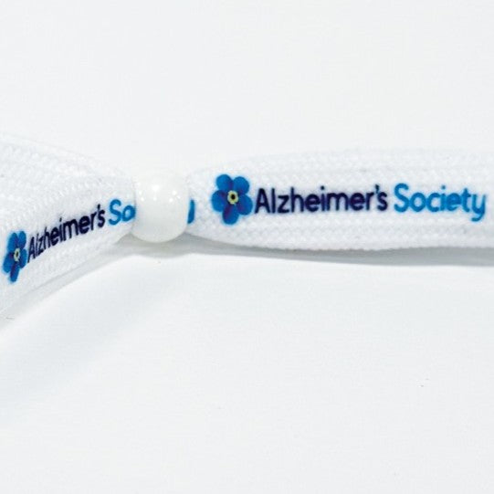 White flat shoelace style fabric wristband in a loop with a white plastic bead sliding closure. Alzheimer's Society logo including the forget-me-not flower is printed several times in full colour on the wristband.