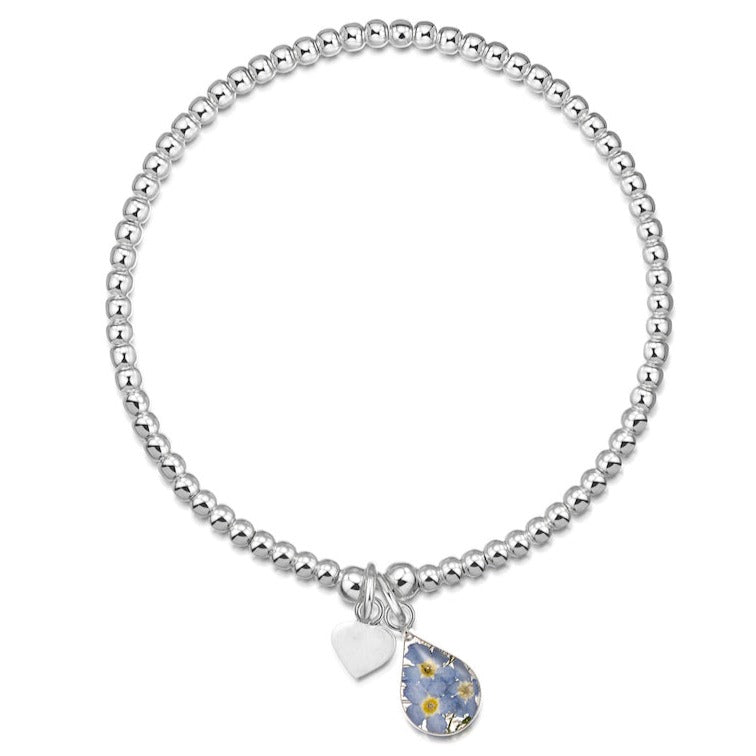 The bracelet is displayed flat to demonstrate its circular shape. It is made up of many small,  round, identical shiny sterling silver beads except for two larger beads positioned next to each other. With the bigger beads hang two charms each attached by a single sterling silver jump ring. The charms are a small, flat silver heart and a larger teardrop of real forget-me-not flowers set in resin and framed in sterling silver. 